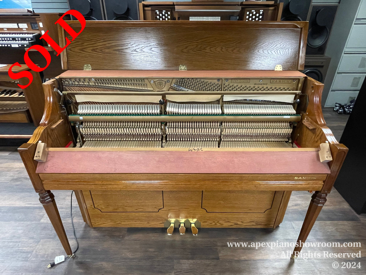 Upright piano with an open top showing internal strings and hammers, placed in a showroom with a wooden finish and a pink felt piano key cover.