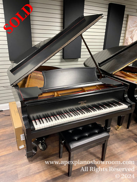 Black grand piano with open lid displaying strings and hammers, keyboard with white and black keys, and a matching piano bench in a showroom setting.