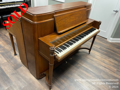 An upright mahogany piano with a bench, situated in a showroom with a herringbone patterned wooden floor.