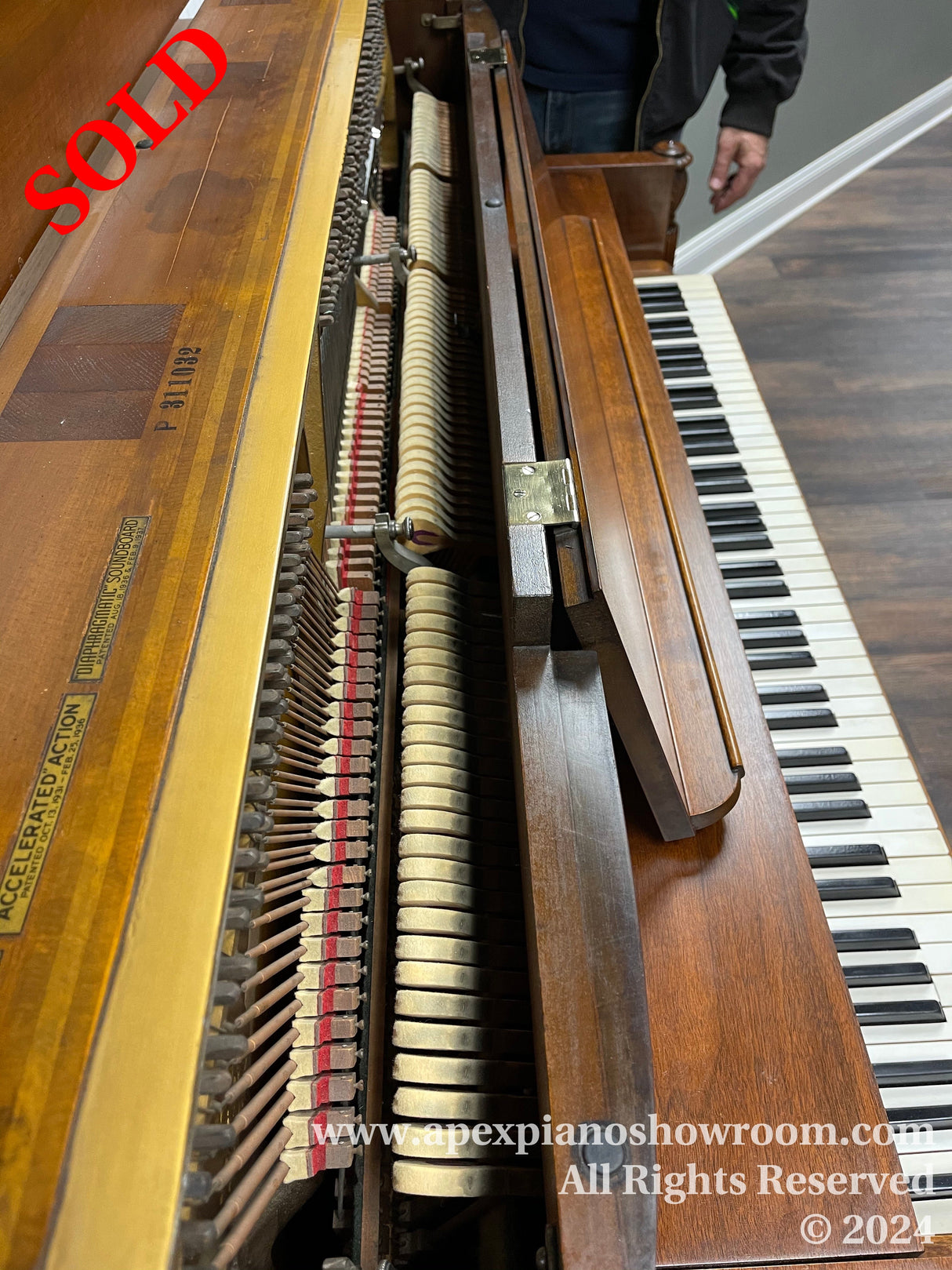 Interior of an upright piano showing the hammers and strings, with the piano’s front panel removed, positioned next to the keyboard, in a showroom setting.