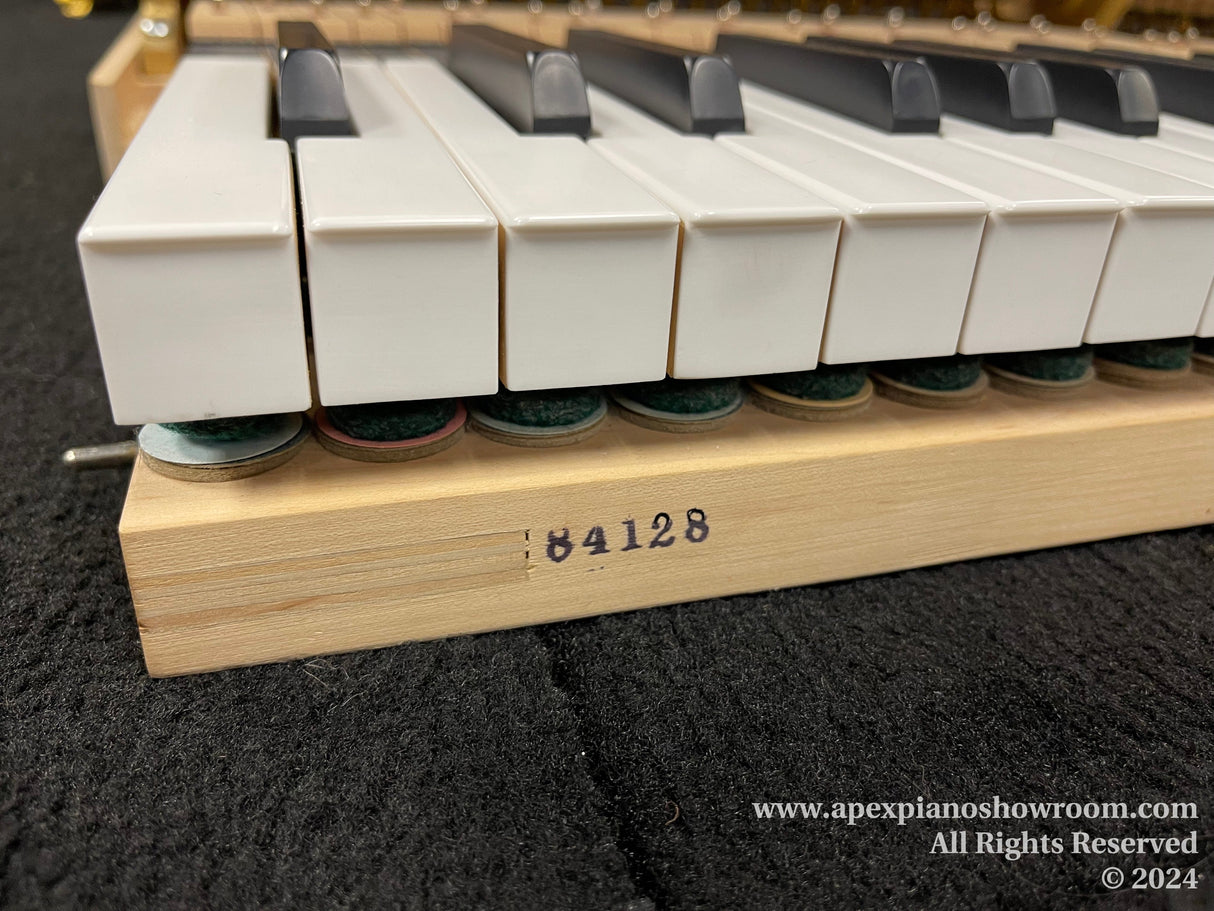 Close-up of piano keys resting on felts above a wooden action rail marked with the number 64128, with a blurred website watermark along the bottom edge.