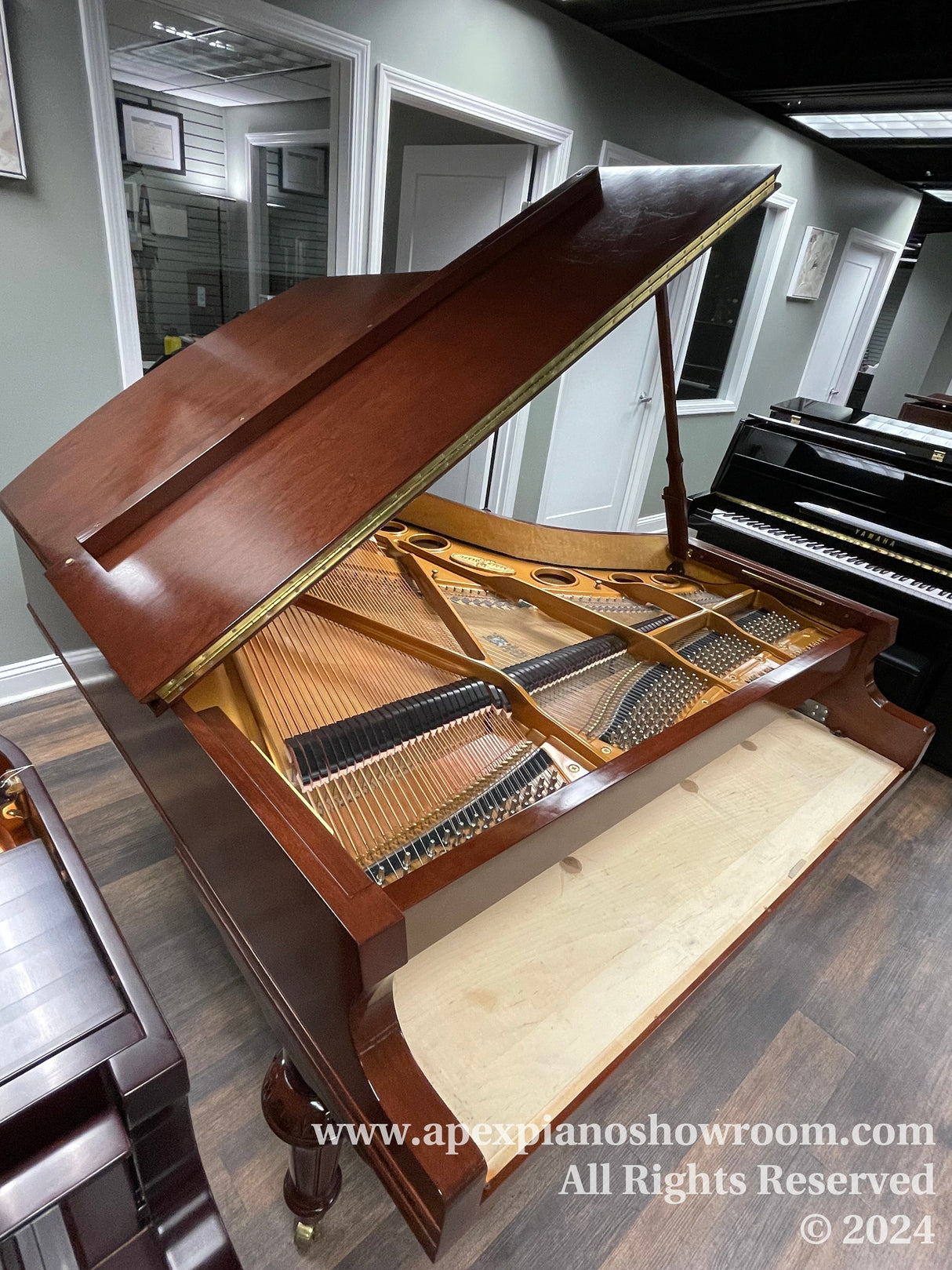 A grand piano with a mahogany finish is open, revealing its intricate internal mechanism of strings and hammers, while the lid is propped up, set against a showroom backdrop with various framed certificates on the wall and a mirror reflecting part of the scene.