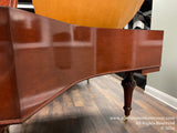 Mahogany grand piano side panel and carved leg detail on showroom floor, reflecting showroom lights, indicating polished wood surface quality.