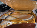 Open grand piano revealing the strings and hammers inside, with a glossy wood finish and brand inscription visible, set in a showroom with other pianos in the background.