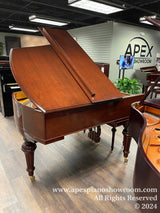 An elegant mahogany grand piano with its lid open, showcasing the detailed interior and strings, standing on a showroom floor with a logo reading APEX SHOWROOM in the background, indicative of a piano retail environment.