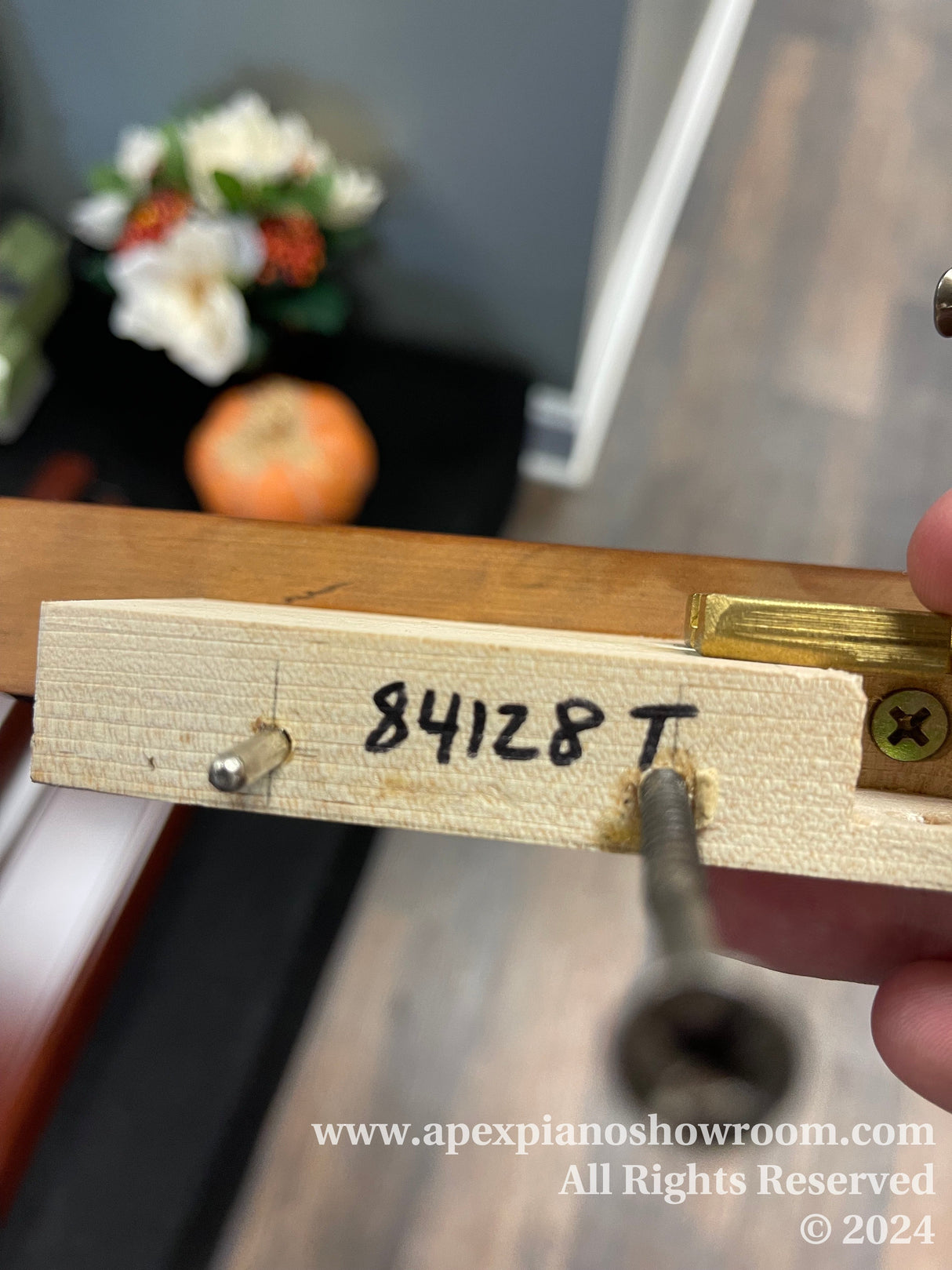 Close-up of a piano part with a serial number 84128T written on it, a tuning pin being inserted, and a blurry background featuring flowers and an orange object.