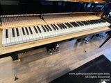 A grand piano with its internal mechanism and keyboard exposed, showcasing the hammers, strings, and keys, model GP-185, in a piano showroom setting.