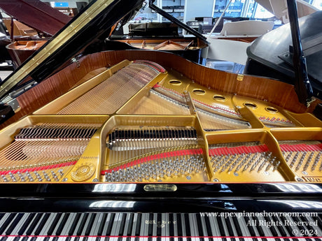 Interior view of a grand piano showing strings and hammers, with the lid open, situated in a showroom with other pianos in the background.