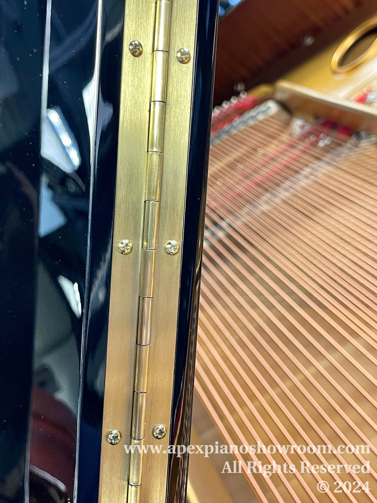 Close-up of a pianos gold-colored hinge on a high-gloss black finish, with strings and tuning pins visible in the background, showcasing the craftsmanship of the pianos hardware and structure.