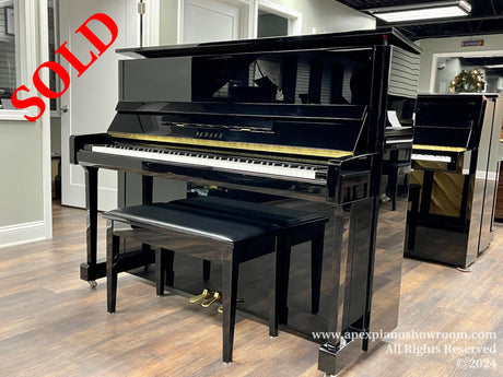 A Yamaha grand piano with a glossy black finish is prominently displayed in a showroom setting, featuring a raised lid revealing its strings and hammers, with a matching piano bench situated in front, set against a backdrop of wood flooring and a softly lit interior.