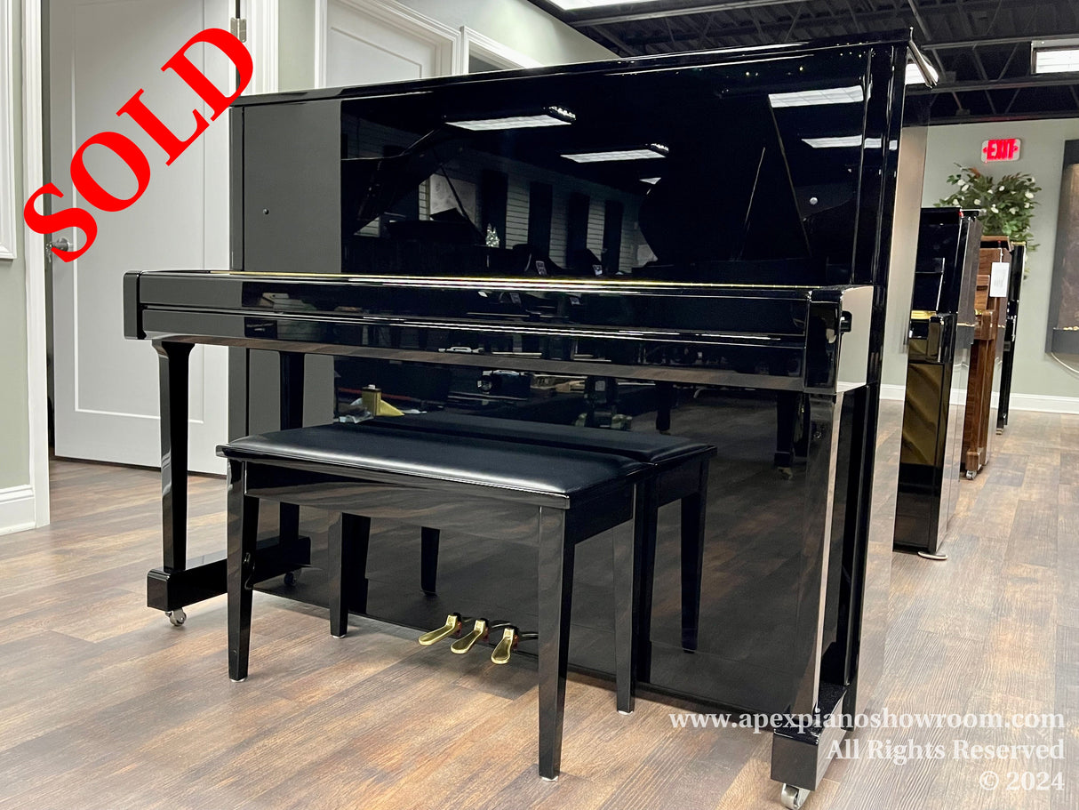 A polished black upright piano with a matching bench on casters, displayed in a showroom with wooden flooring, surrounded by other pianos in the background, reflecting the interior lighting and space.