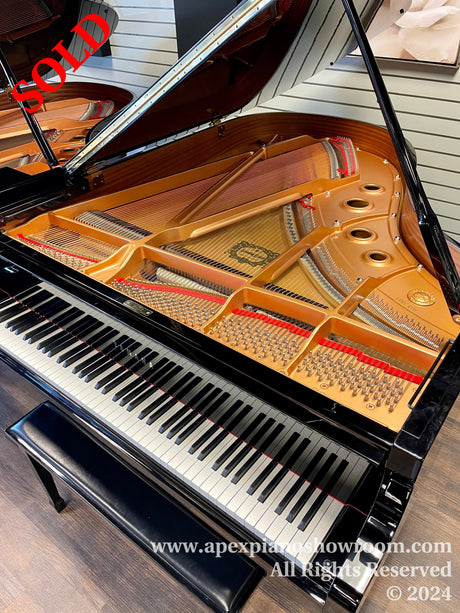 A grand piano with its lid open, showing the intricate internal strings and hammers, accompanied by a matching piano bench, set in a well-lit showroom with wood flooring.