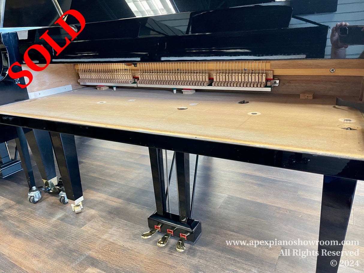 Grand piano with its action removed, showing the empty interior with hammers and strings visible, set against a wooden bench in a showroom setting.