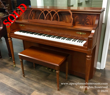 A mahogany Yamaha upright piano with intricate wood carving on the front panel and matching stool, set against a showrooms hardwood floor.
