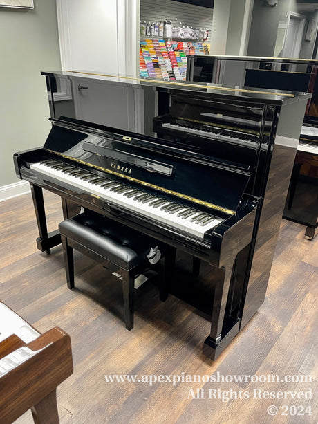 A Yamaha upright piano with a glossy black finish and matching bench placed on a wooden floor in a well-lit showroom with sheet music displayed in the background.