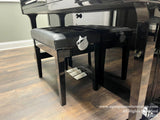 Black grand piano with matching bench on a wooden floor in a showroom setting.