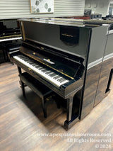 A glossy black Yamaha upright piano situated in a showroom with a wooden floor and a light-colored wall with a framed picture in the background.