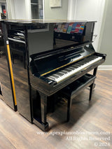 A glossy black upright piano with an open lid revealing the keyboard, set against a showroom backdrop with music books in the background, positioned on a wooden floor.
