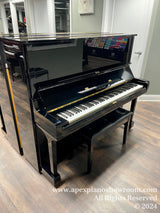 A glossy black upright Yamaha piano with the lid closed, situated in a well-lit showroom with wooden flooring and decorative elements reflected on its polished surface.