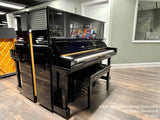 A black grand piano on display in a showroom with a glossy finish and the lid open, sitting on a brown hardwood floor with music stands and other pianos visible in the background.