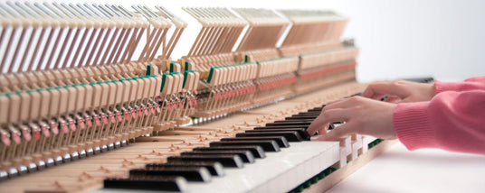 A person's hands playing a grand piano with the interior mechanism exposed, showcasing the hammers and strings.