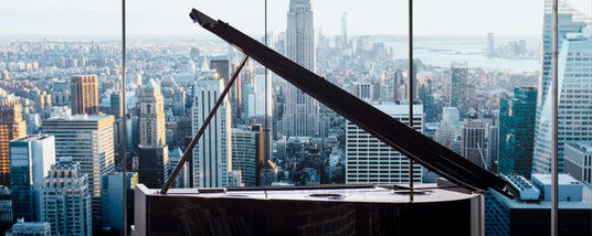 Grand piano with a glossy finish positioned by a large window overlooking a cityscape, symbolizing a fusion of urban architecture and classical music elegance.