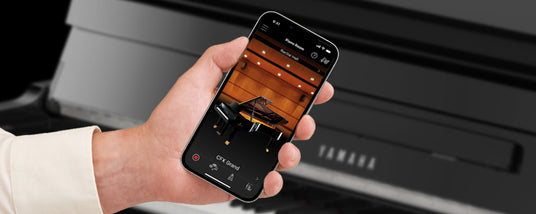 Person holding a smartphone with an app displaying a grand piano, in front of a digital piano keyboard.