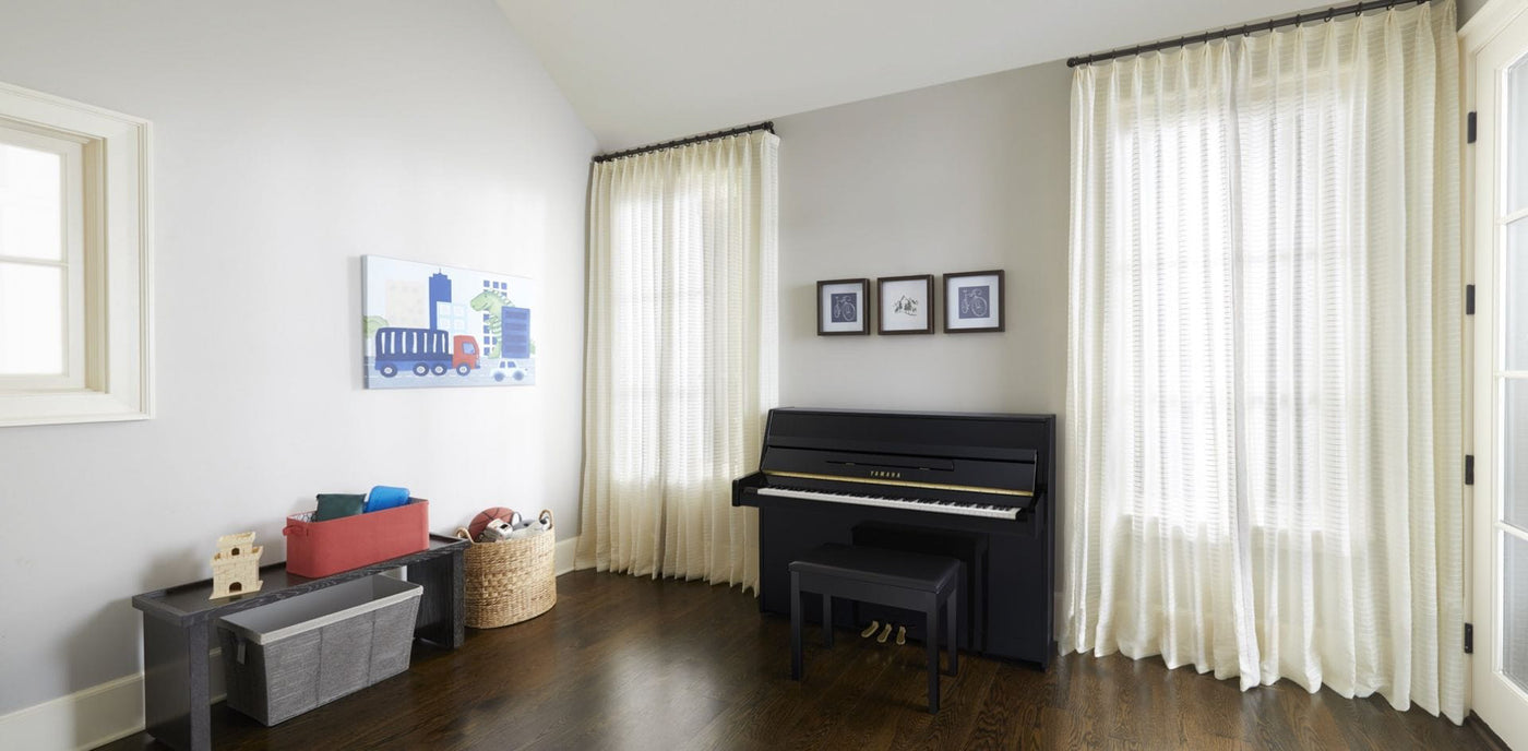 A modern upright black piano with a matching bench in a bright room with wooden floors, white curtains, and decorative artwork, creating an elegant space for music practice or teaching.