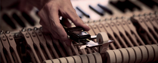 Piano technician tuning a grand piano by adjusting the tension of the strings using a tuning lever, with the piano's action partially exposed.