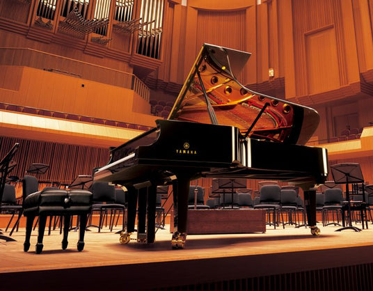 A grand Yamaha piano on stage in a concert hall with rows of empty chairs and music stands, indicating a rehearsal or performance setting in the piano industry.