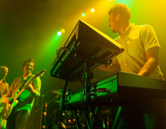 Keyboard player performing on stage with an electronic piano at a concert with band members and green stage lighting in the background.