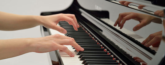 A person's hands playing a piano, with one hand reflected in the piano's surface.