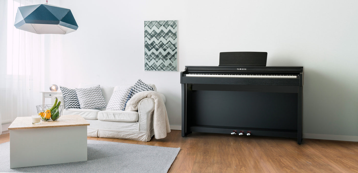 A modern living room featuring a Yamaha digital piano with a full keyboard, set against a white wall adjacent to a cozy couch, with a geometric-patterned artwork above, illustrating a contemporary home music setup.