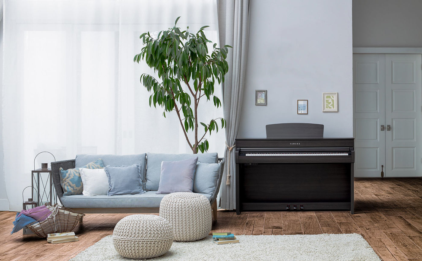 A modern living room with a dark wood Yamaha digital piano near a white wall, beside a large potted plant and a comfortable sofa with cushions.