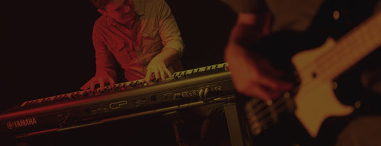 Musician playing a Yamaha electric piano on stage with a guitarist in the background, set in a dimly lit concert setting.