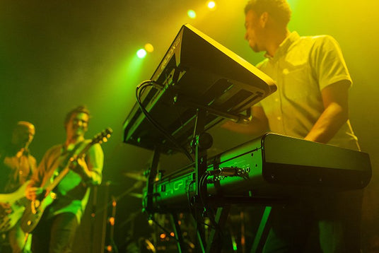 Keyboard player performing on stage with digital piano in a live concert setting, flanked by a guitarist and illuminated by green stage lights.