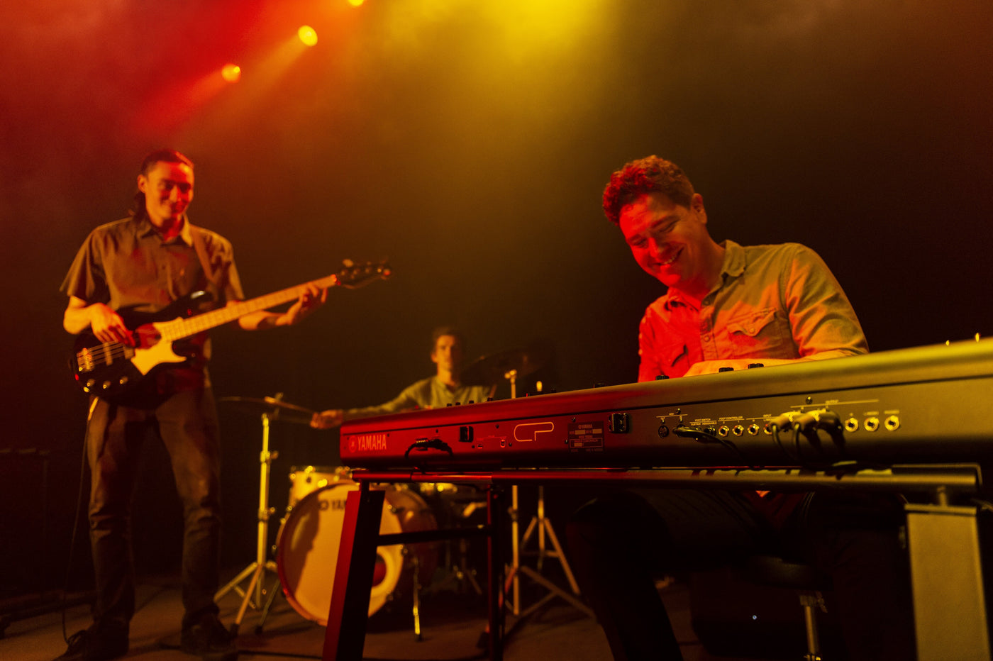 Electronic stage piano being played by a musician on stage with warm lighting, with other band members in the background including a bass guitarist and a drummer.