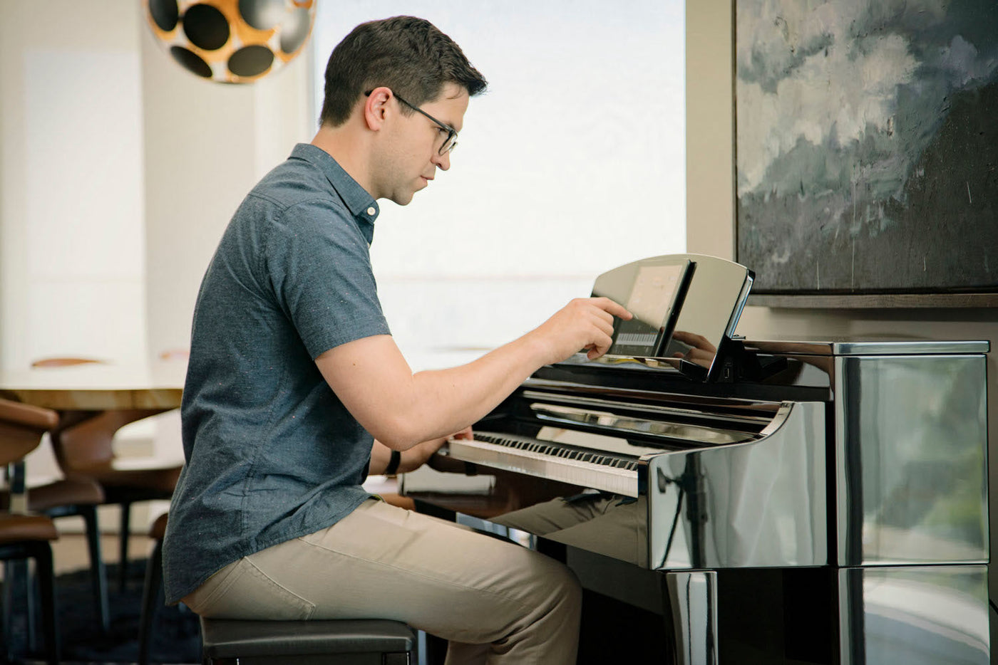 Man practicing piano using a digital tablet for sheet music, seated at a sleek, modern black piano in a well-lit room with interior decor.