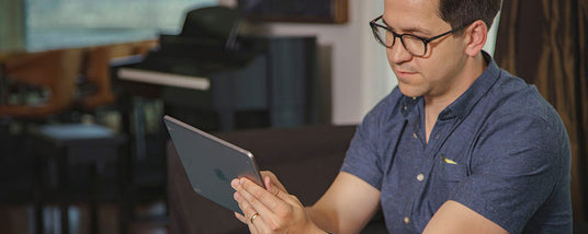 Man in glasses and blue shirt using a tablet while sitting on a couch in a living room.