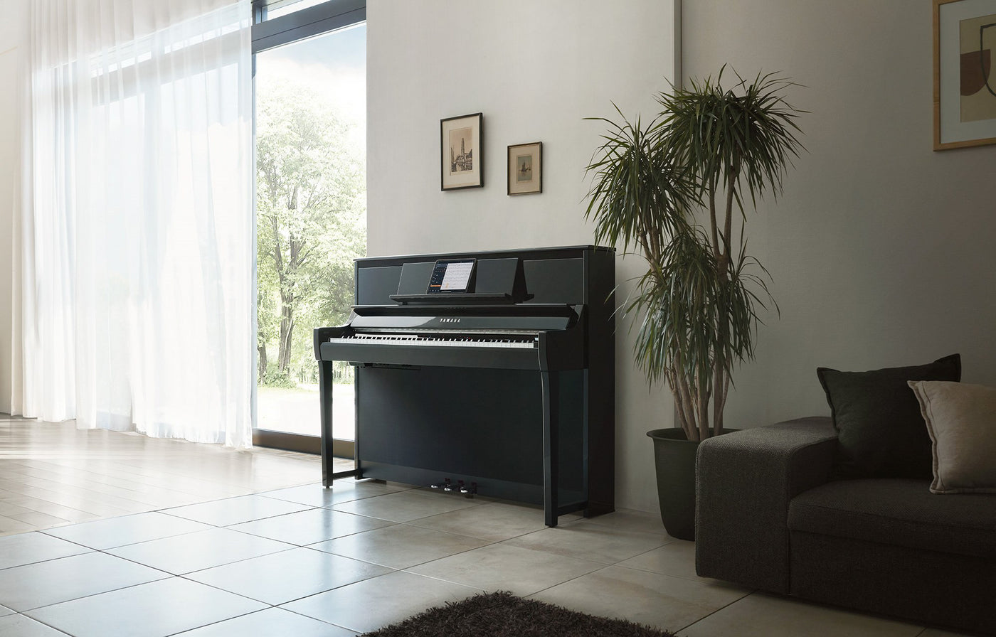 A modern digital piano in a bright living room with large windows, set against a matte wall, adjacent to a plush sofa and an indoor potted plant.