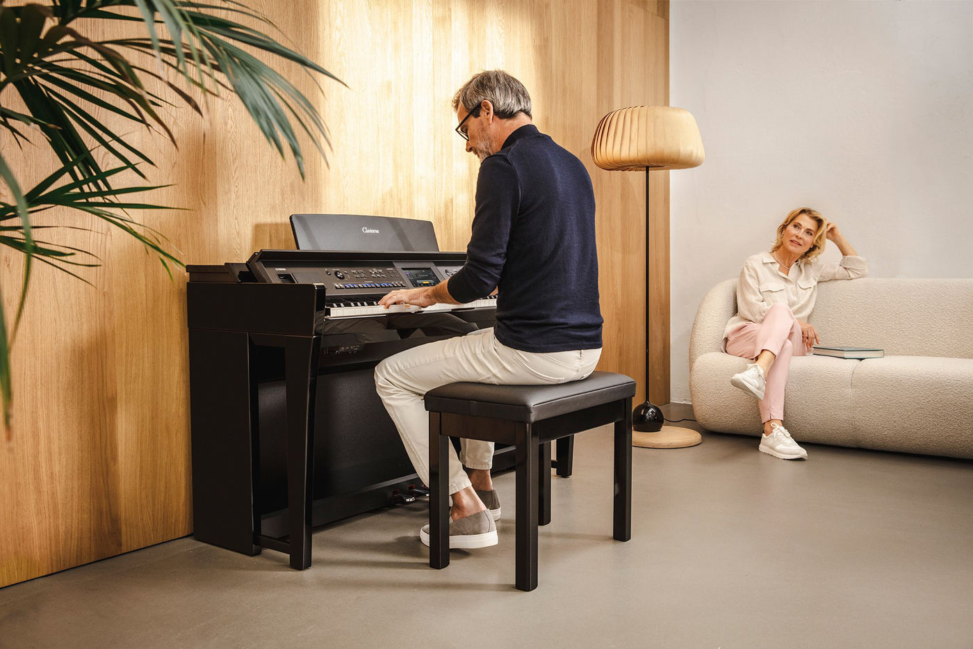 A man playing a modern digital piano with a woman sitting on a couch listening in a well-lit living room with wooden flooring and a warm ambiance.