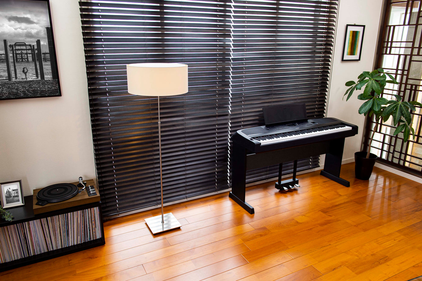 modern living room with a digital piano next to a window with horizontal blinds, a floor lamp, a turntable on a cabinet filled with vinyl records, framed pictures, and a potted plant.