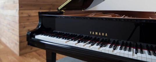 Side view of a black Yamaha grand piano with open lid, showcasing the keyboard and the brand name, placed in a room with wooden flooring and paneling.