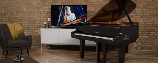 Black grand piano positioned in a cozy living room setting, with a television displaying a pianist in the background.