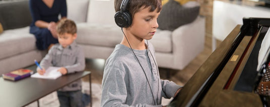 Young boy practicing piano using headphones with another child doing homework at the table and an adult supervising in a home setting, showcasing the versatility of modern pianos in providing silent practice options.