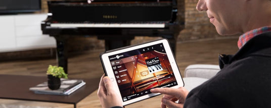 Person using a tablet with piano app in a modern living room with an acoustic piano in the background.
