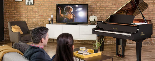 A Yamaha grand piano in a cozy living room setup, with a couple watching a concert on the television, potentially drawing inspiration for piano playing.