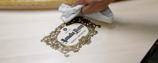 Close-up of a person's hand polishing a grand piano's nameplate that reads Amanda Divino with an ornate design, representing the final touches in piano craftsmanship.