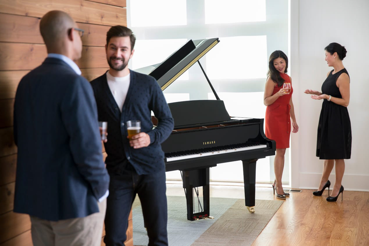 Group of people socializing around a Yamaha grand piano in a modern home setting, suggestive of a high-end piano showroom or a music-related gathering.