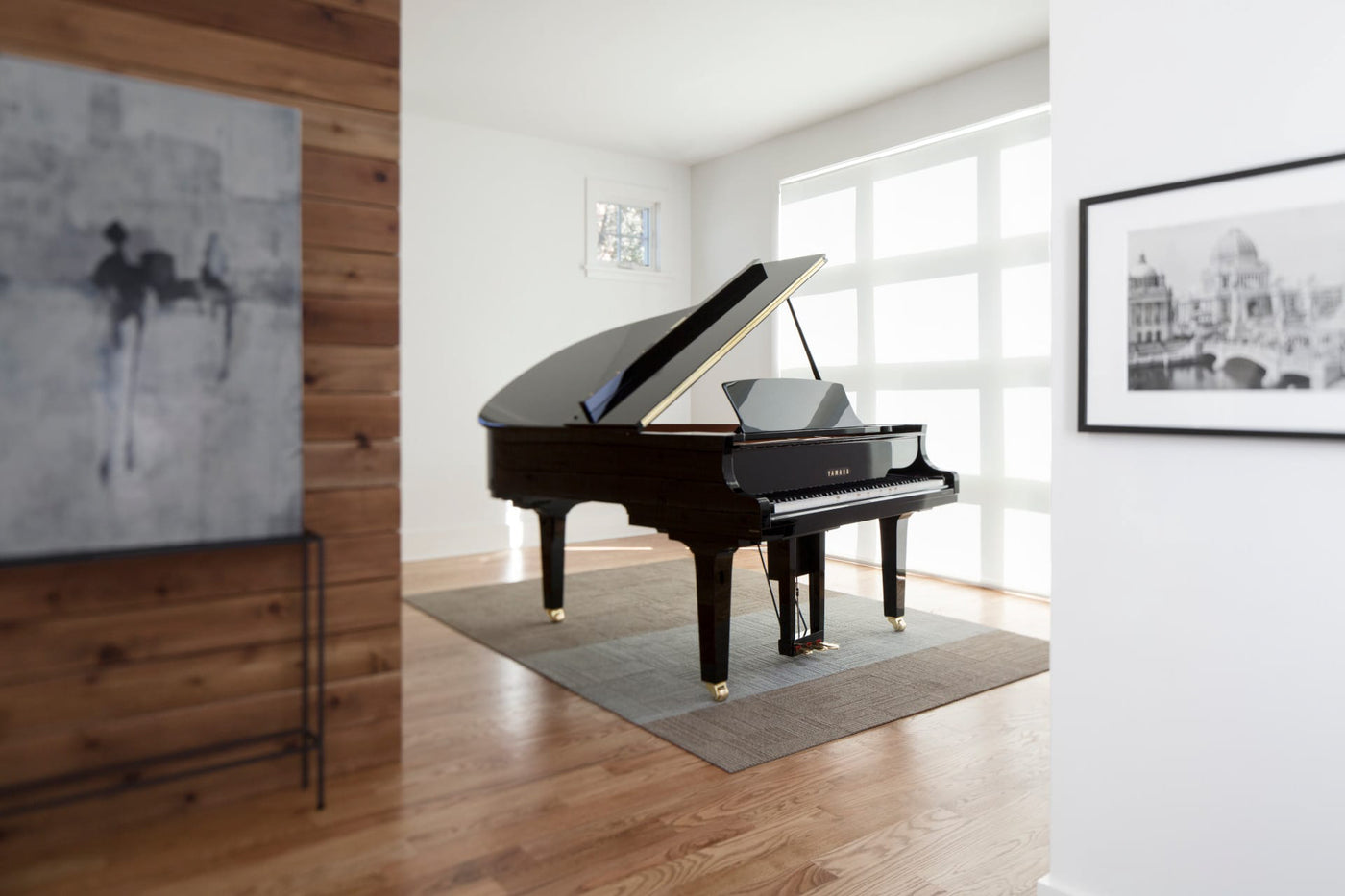 Elegant black grand piano positioned in a bright room with wood flooring, artwork on the walls, and large windows providing natural light.